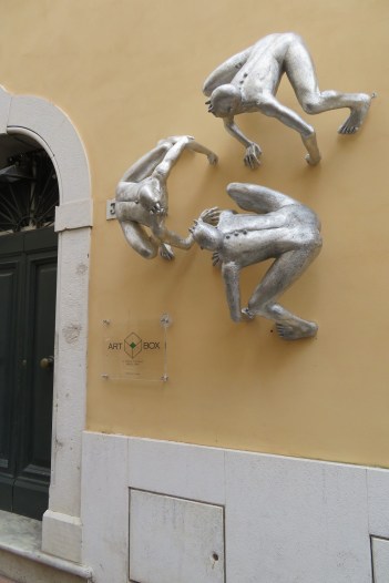 Three silver figures crouch on a wall in Pietrasanta
