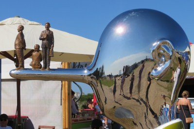 A shiny man's head with a long pointy nose and three people are standing on the nose